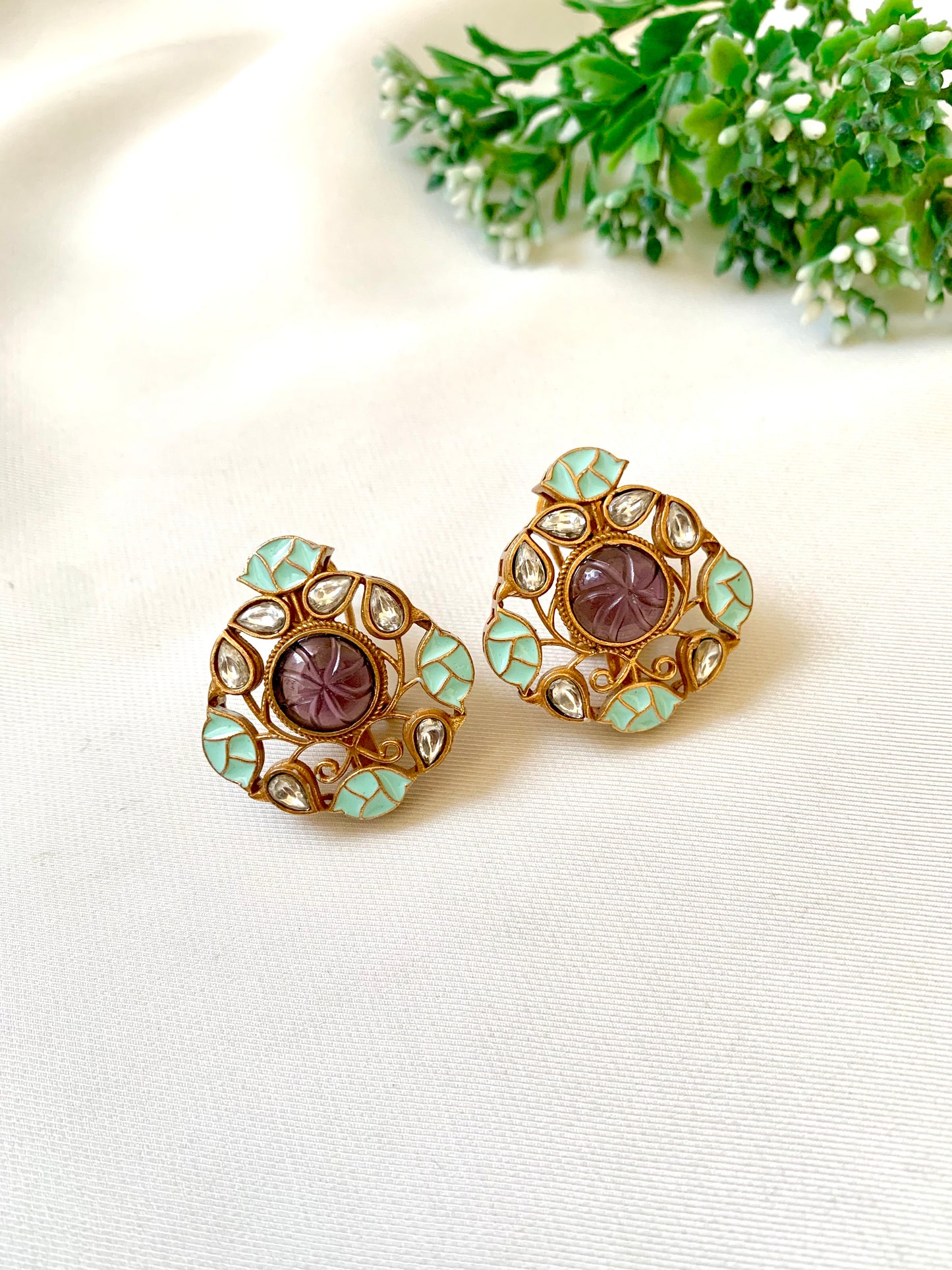 A pair of gold-colored stud earrings featuring amethyst stones and green accent stones, surrounded by intricate metalwork.