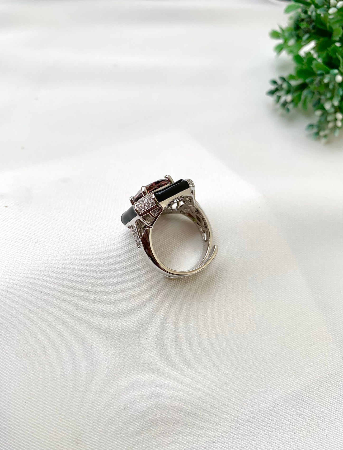 A silver-toned ring with a central black rectangular stone and small clear stones, displayed on a white textured fabric surface with a small green plant in the corner.