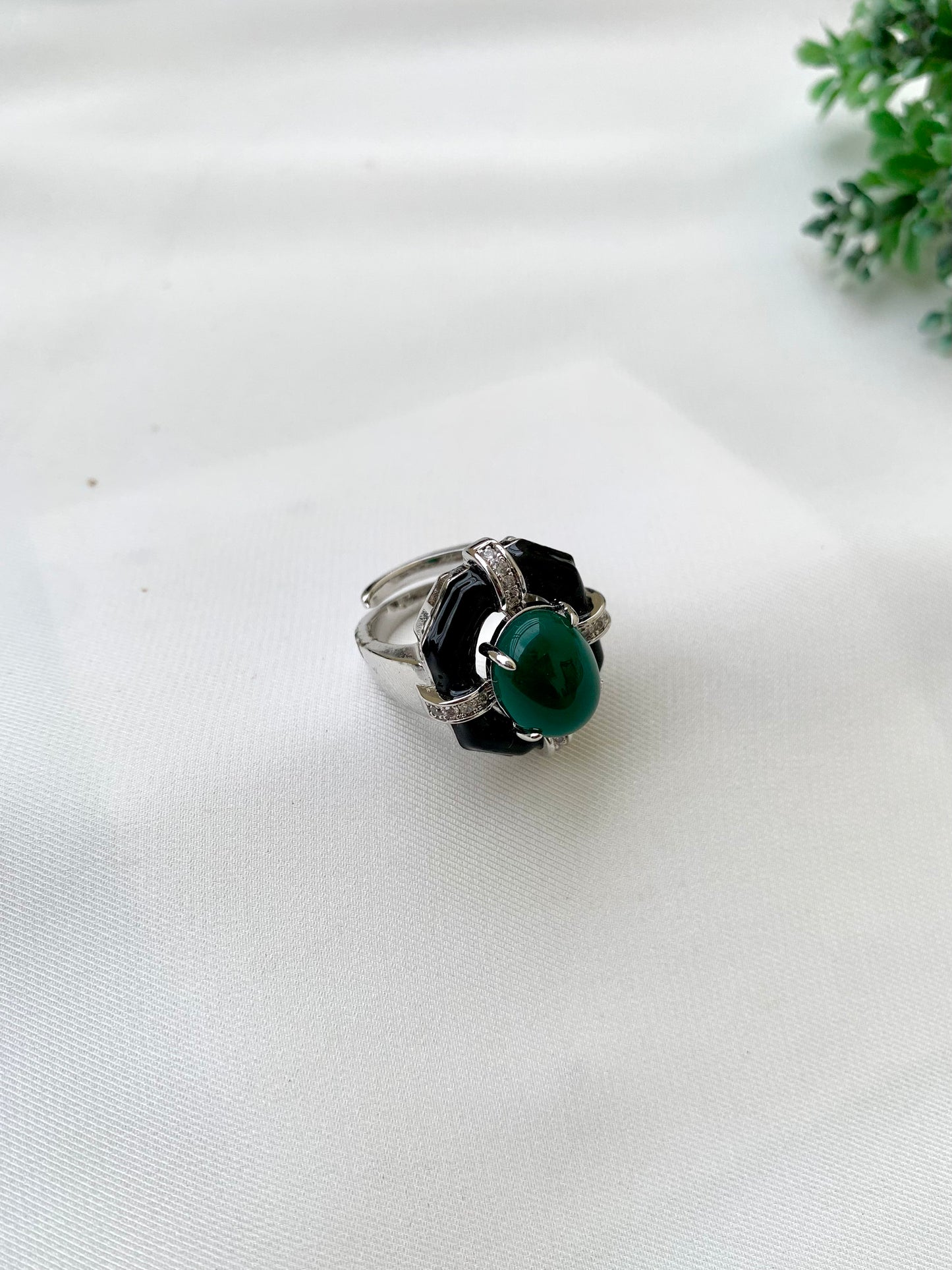 A silver-toned ring with a large oval green cabochon stone and black petal-like accents, photographed on a white fabric surface with a small green plant in the corner.
