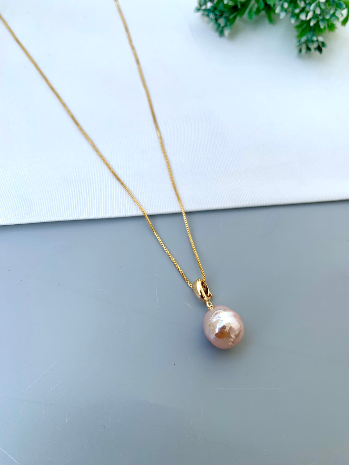 A gold chain necklace with a single baroque-style pink pearl pendant, displayed on a split white and gray surface with a small green plant in the corner.