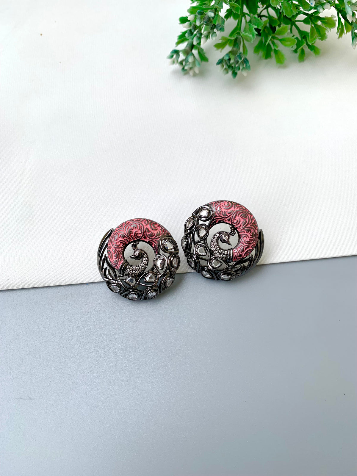 A pair of round, ornate earrings with a dark metallic filigree border and a pink and silver spiral center, displayed on a split white and gray surface with a small green plant in the corner.