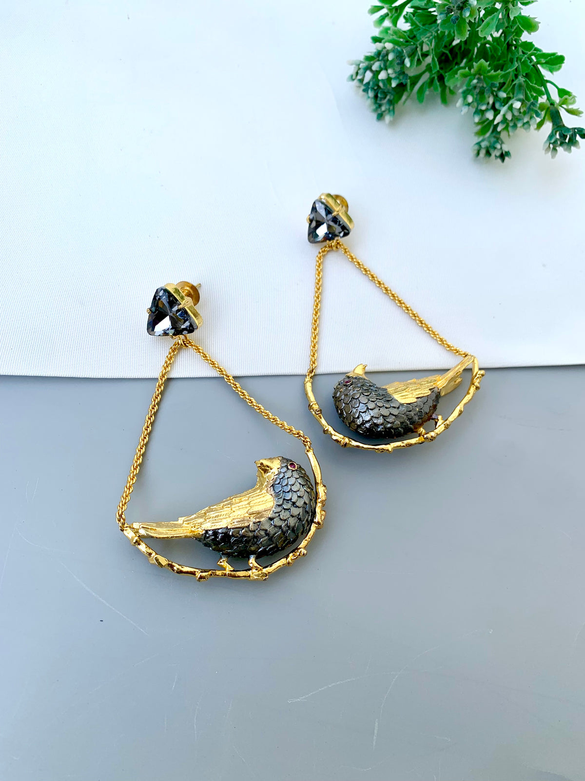 A pair of gold and black bird drop earrings with textured scales, displayed on a split white and gray surface with a small green plant in the corner.
