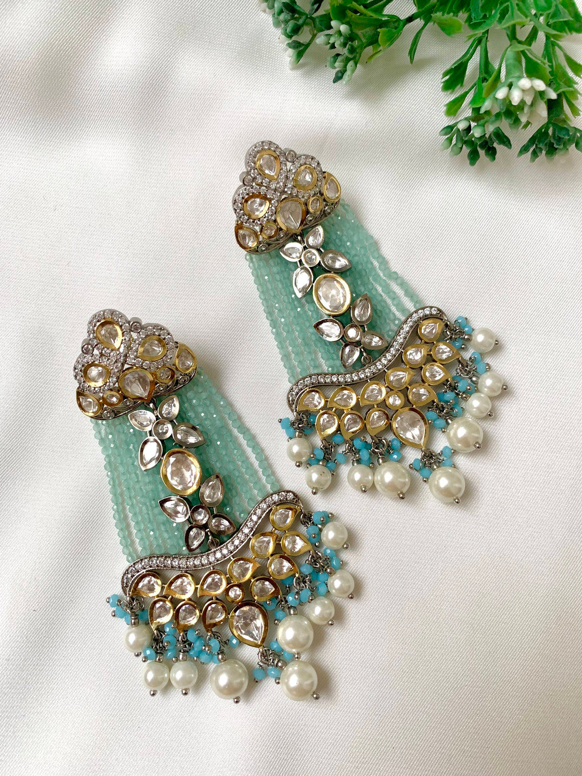 A pair of ornate gold-toned jhumka earrings with mint green beaded tassels, decorated with white faux pearls, clear crystals, and turquoise blue beads, displayed on a white fabric background with green leaves in the corner.