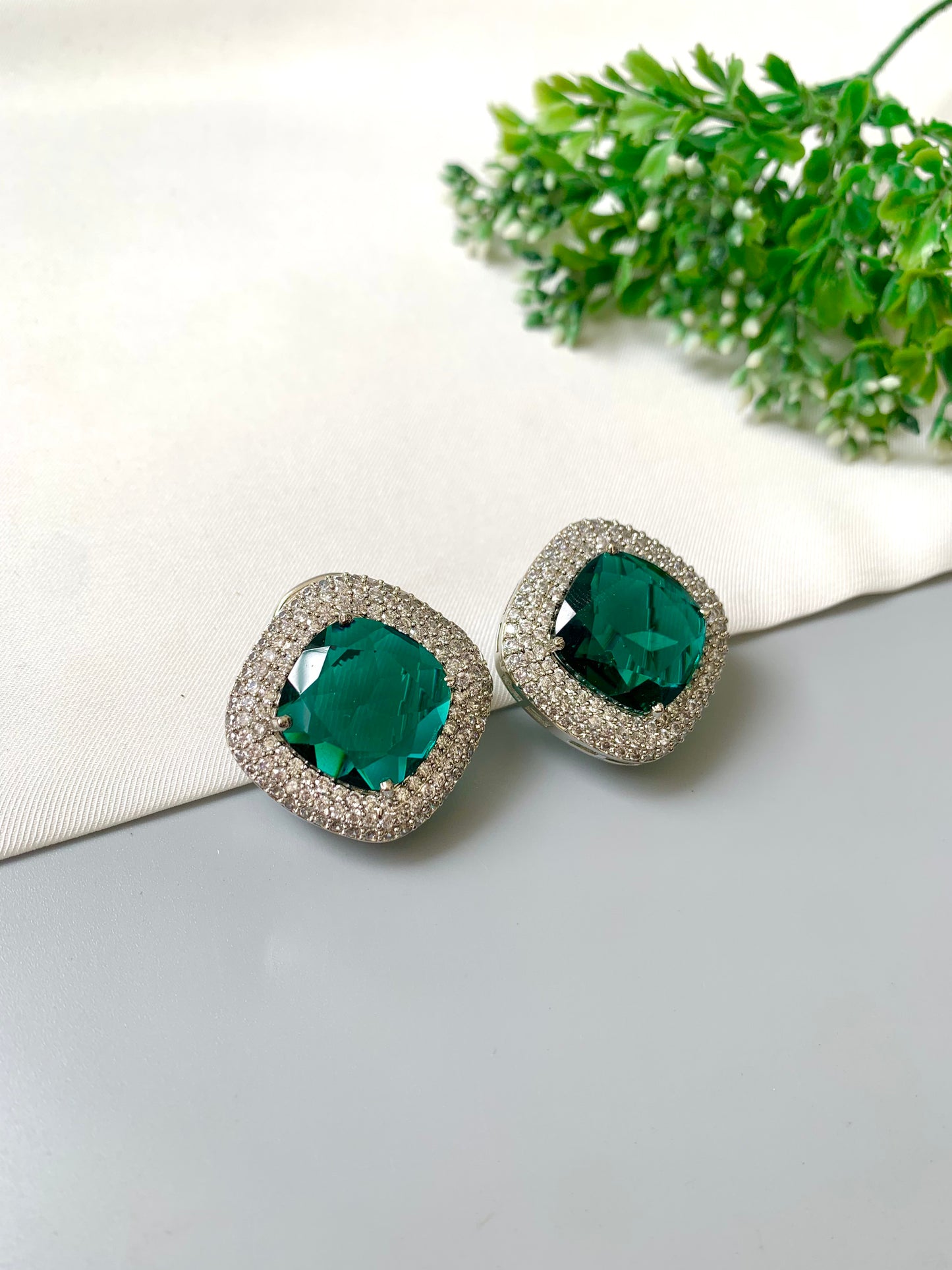 Pair of emerald green earrings with silver accents on a light surface with green leaves in the background.