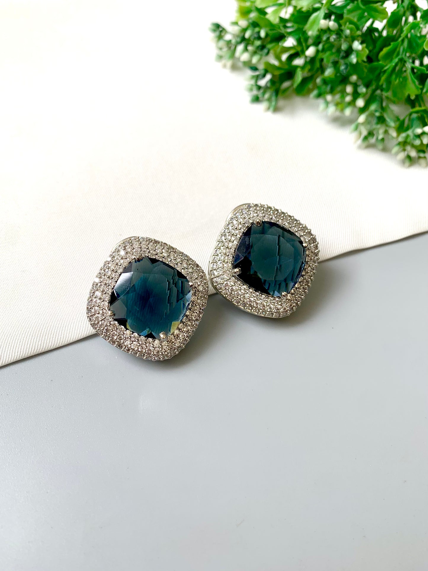 Pair of earrings with blue gemstones on a light surface with greenery in the background