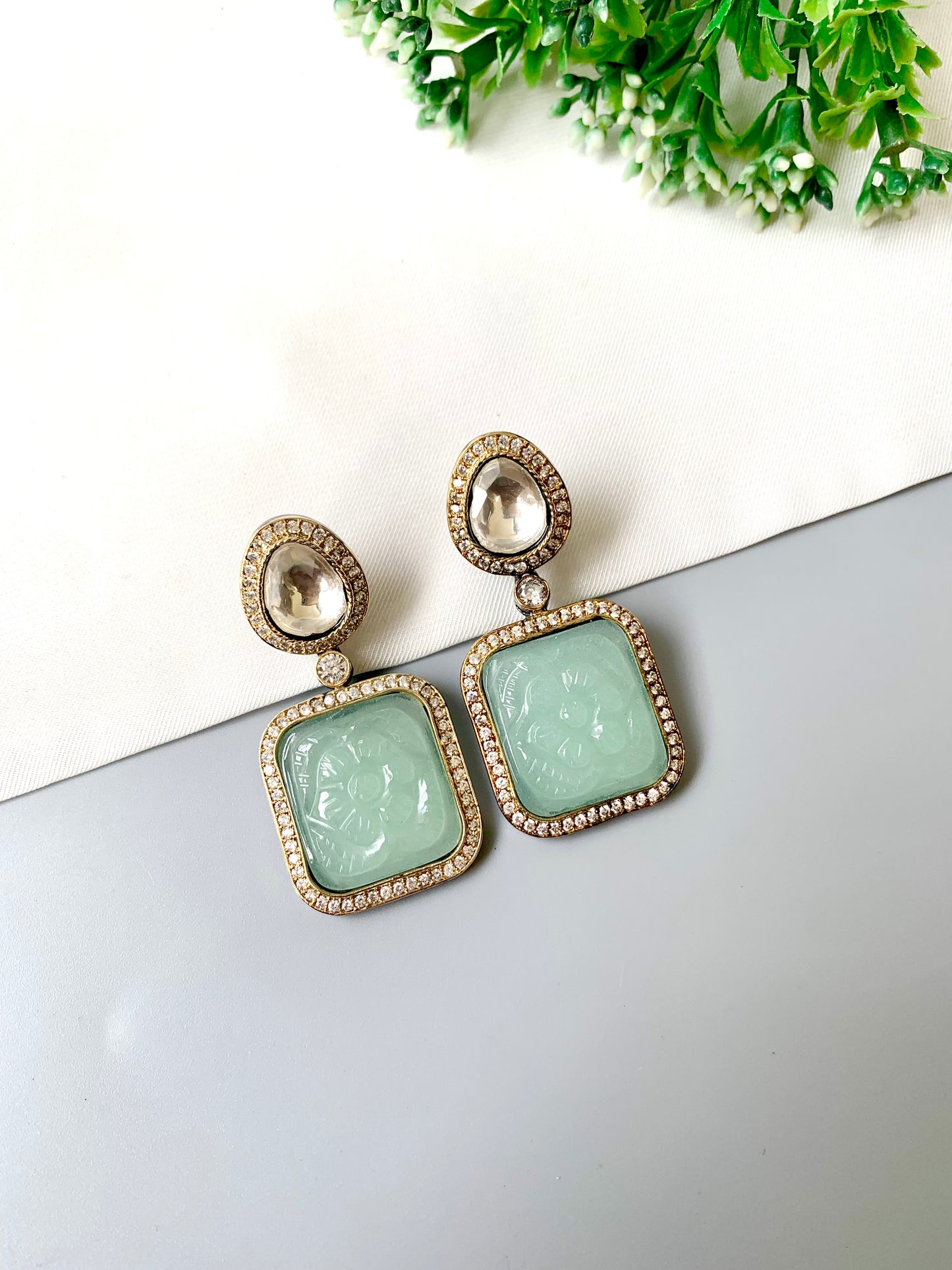 Pair of earrings with green gemstones on a white surface with greenery in the background