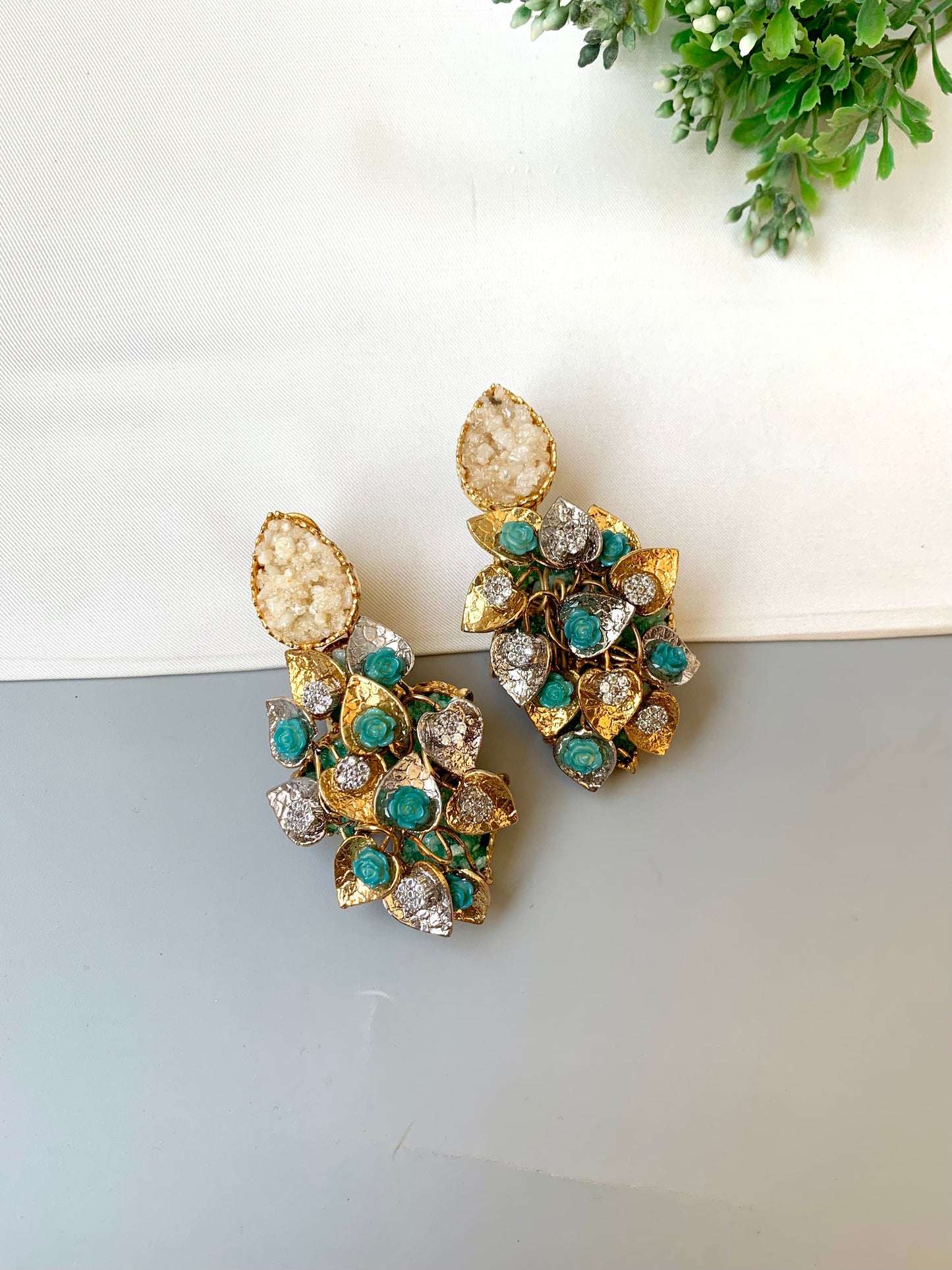 Gold and green gemstone earrings on a light gray surface with a white and green plant in the background.