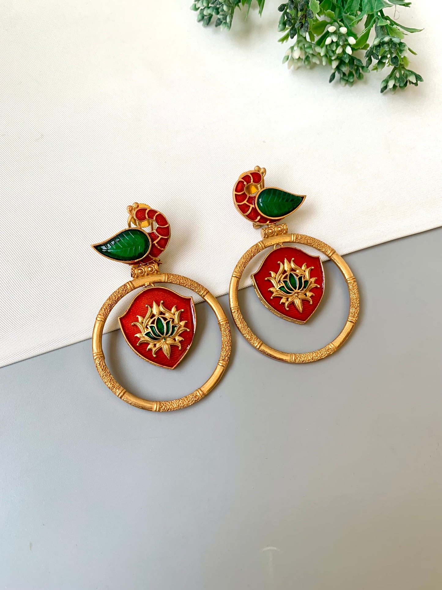 Pair of earrings with red, green, and gold design on a white surface with a plant in the background.