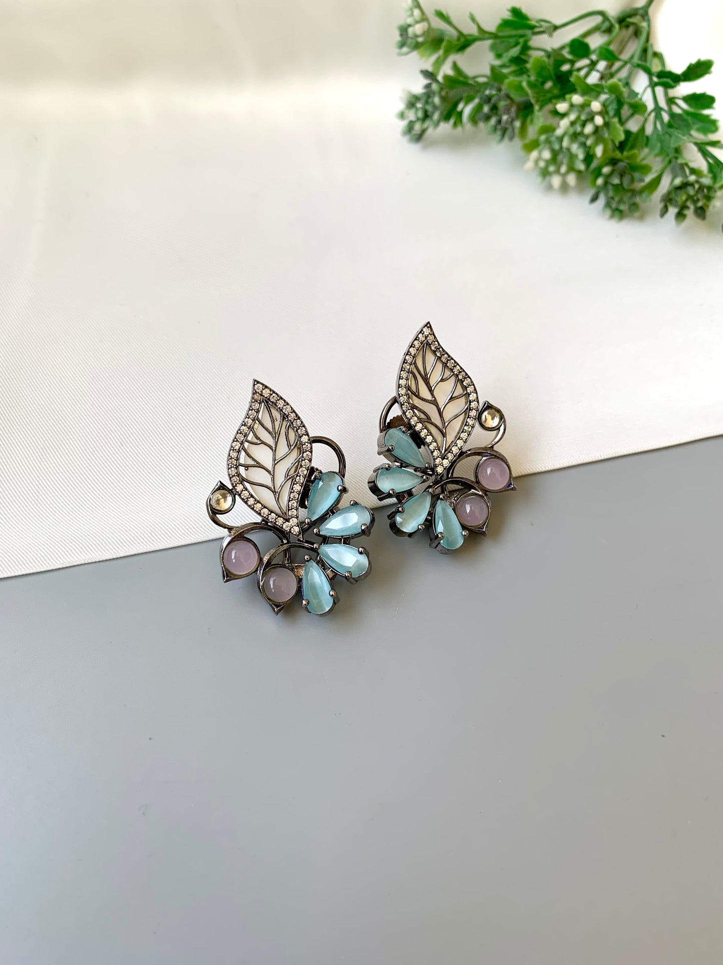 Pair of decorative earrings with leaf design on a light surface with greenery in the corner.