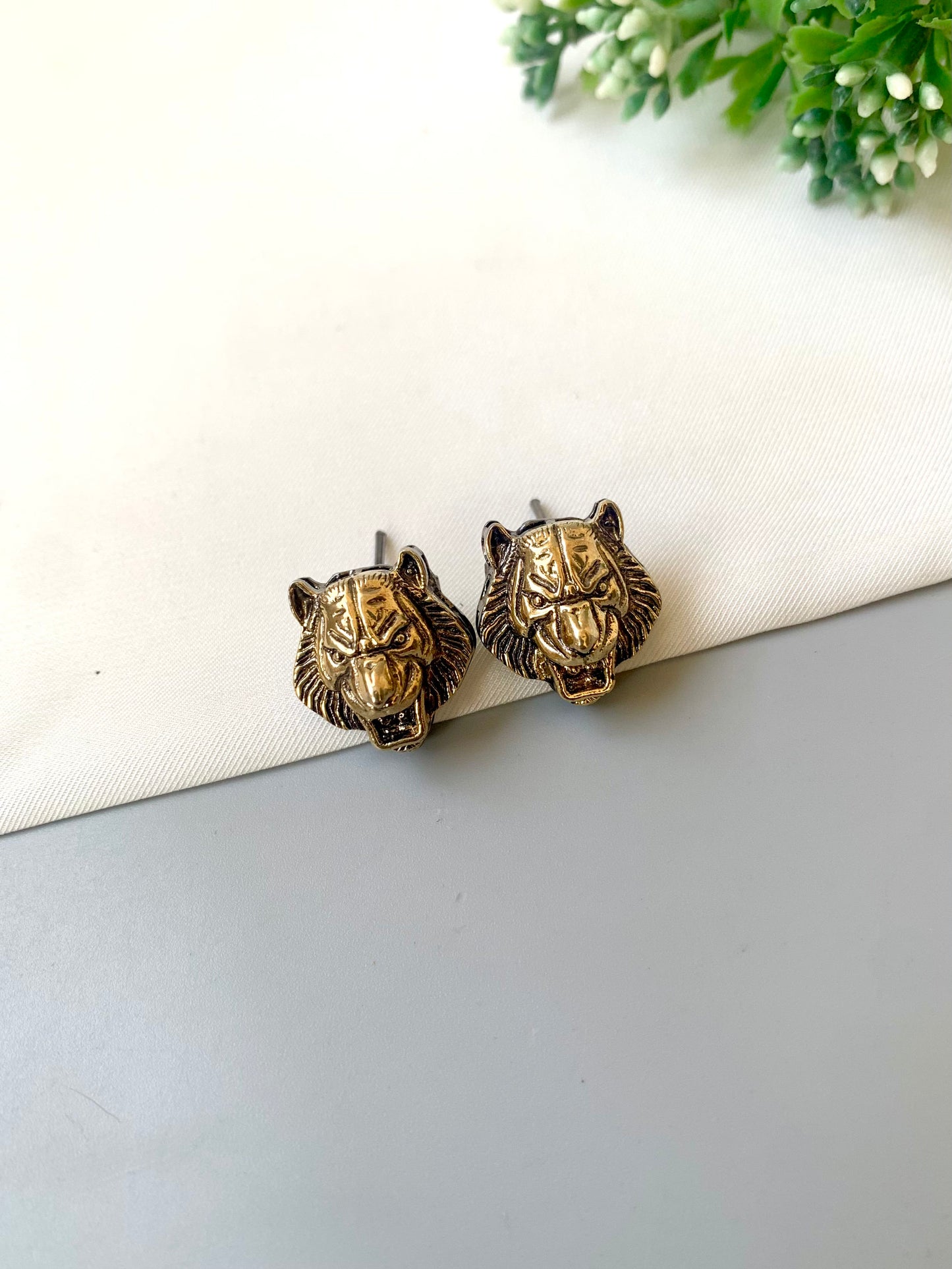 Pair of gold lion head earrings on a white surface with greenery in the background