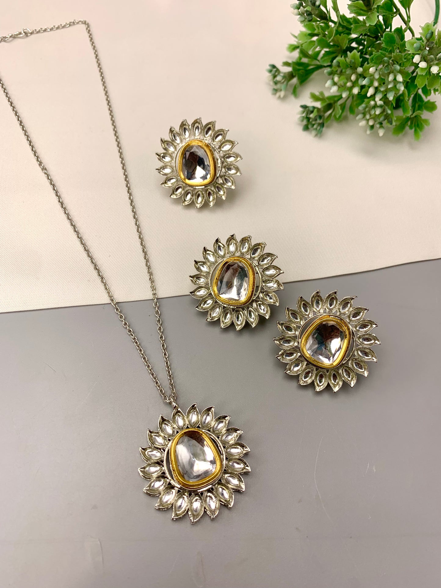 A jewelry set featuring a necklace and three matching earrings with silver-toned floral sunburst settings, clear rhinestone accents, and oval clear gemstones, arranged on a split cream and gray surface with a small green plant in the corner.