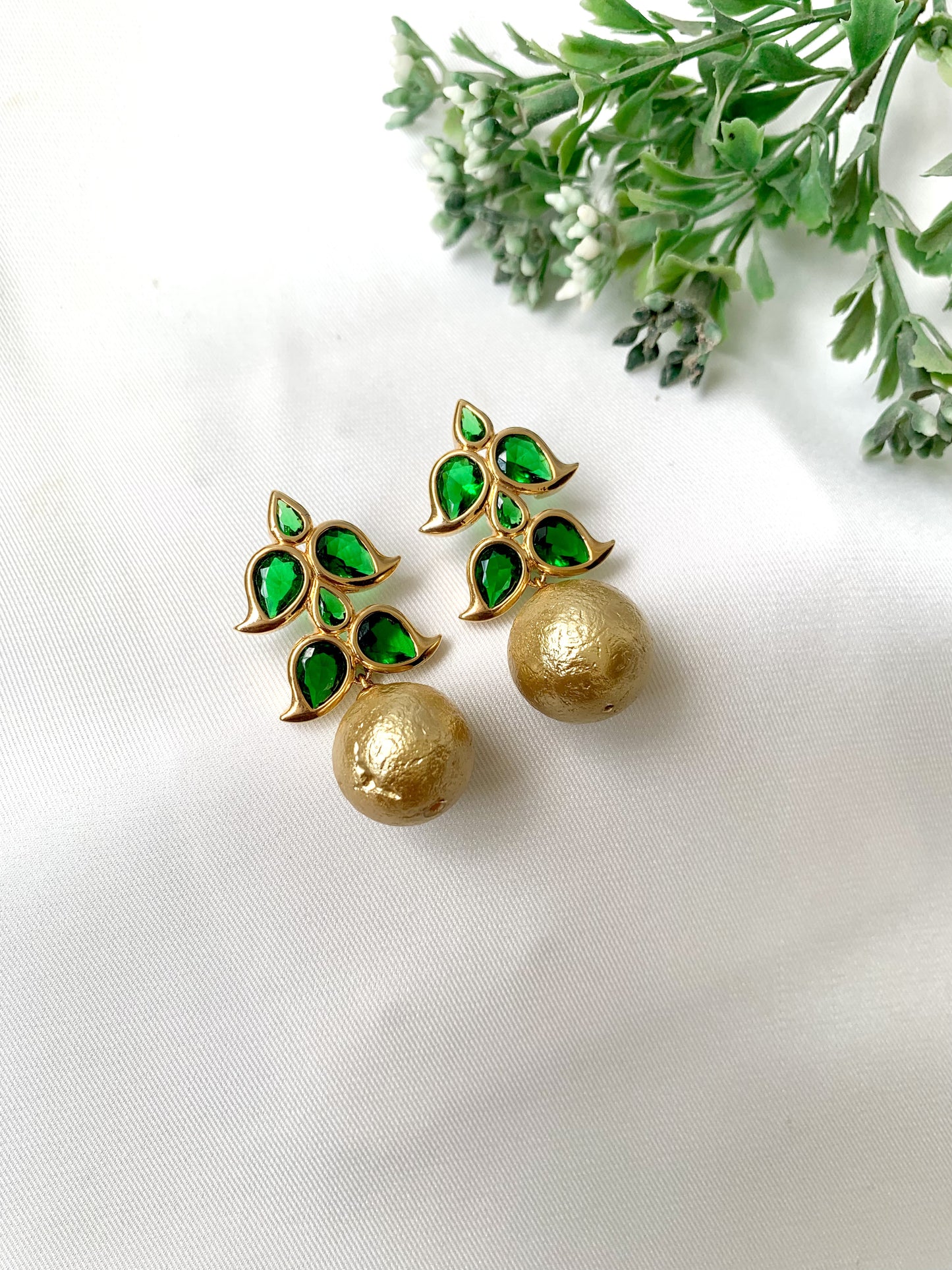 Gold earrings with green gemstones on a white background with greenery.