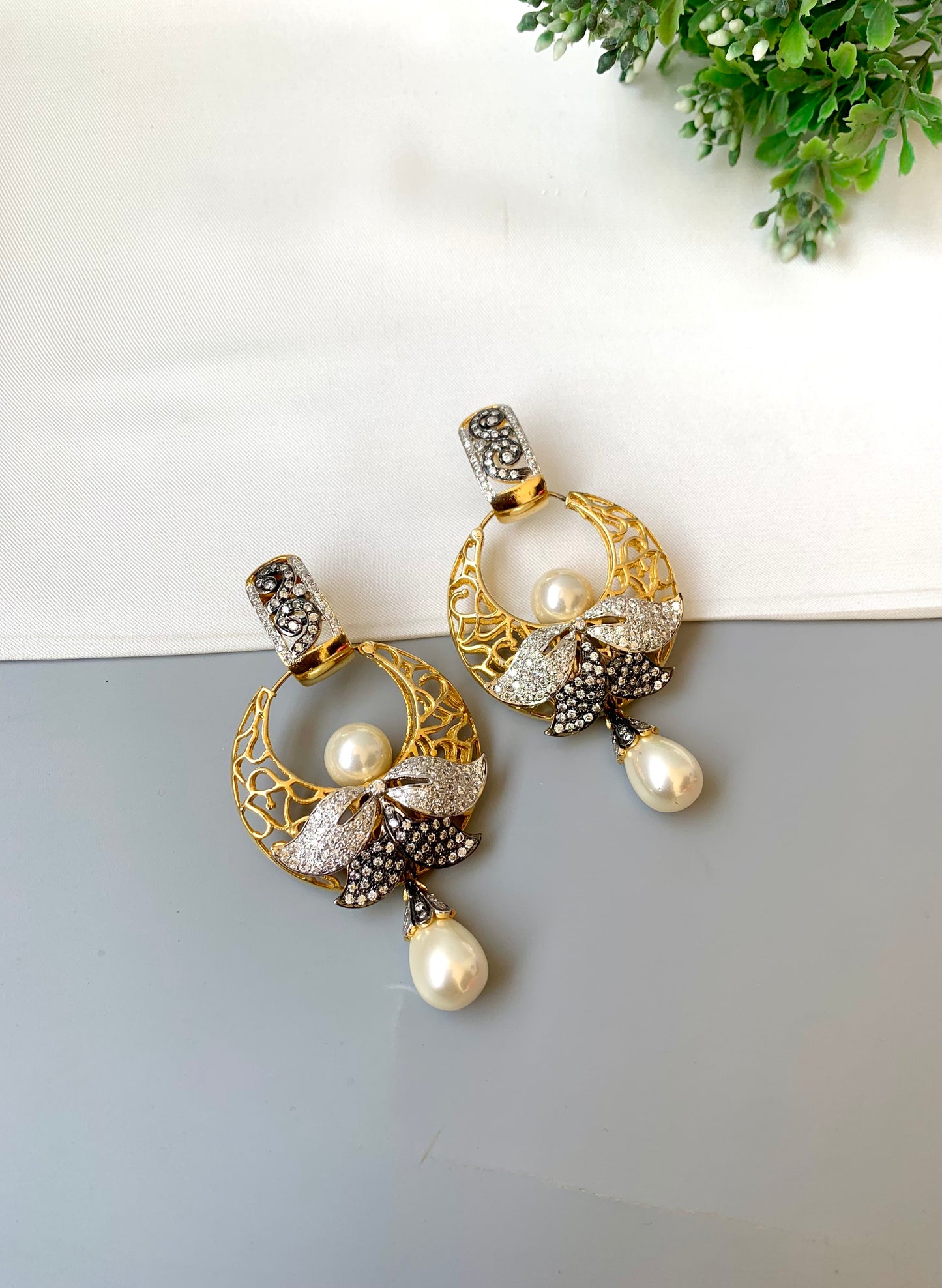 Gold and pearl earrings on a light gray surface with a green plant in the background