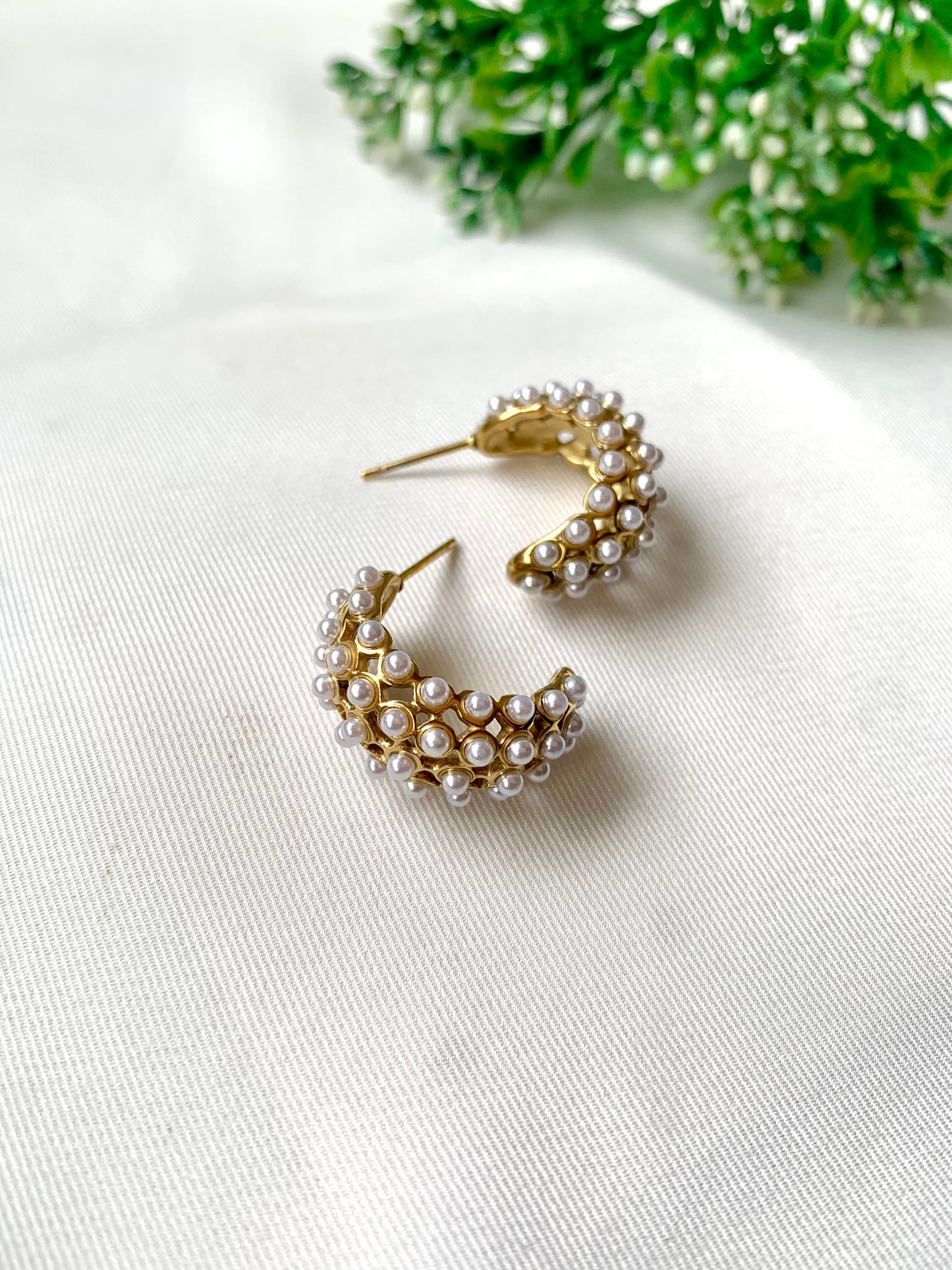Gold hoop earrings with pearl accents on a white surface with greenery in the background