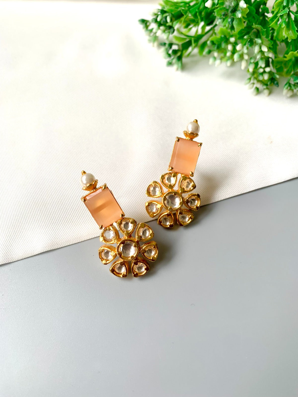 Gold earrings with peach-colored stones on a light gray surface with greenery in the background
