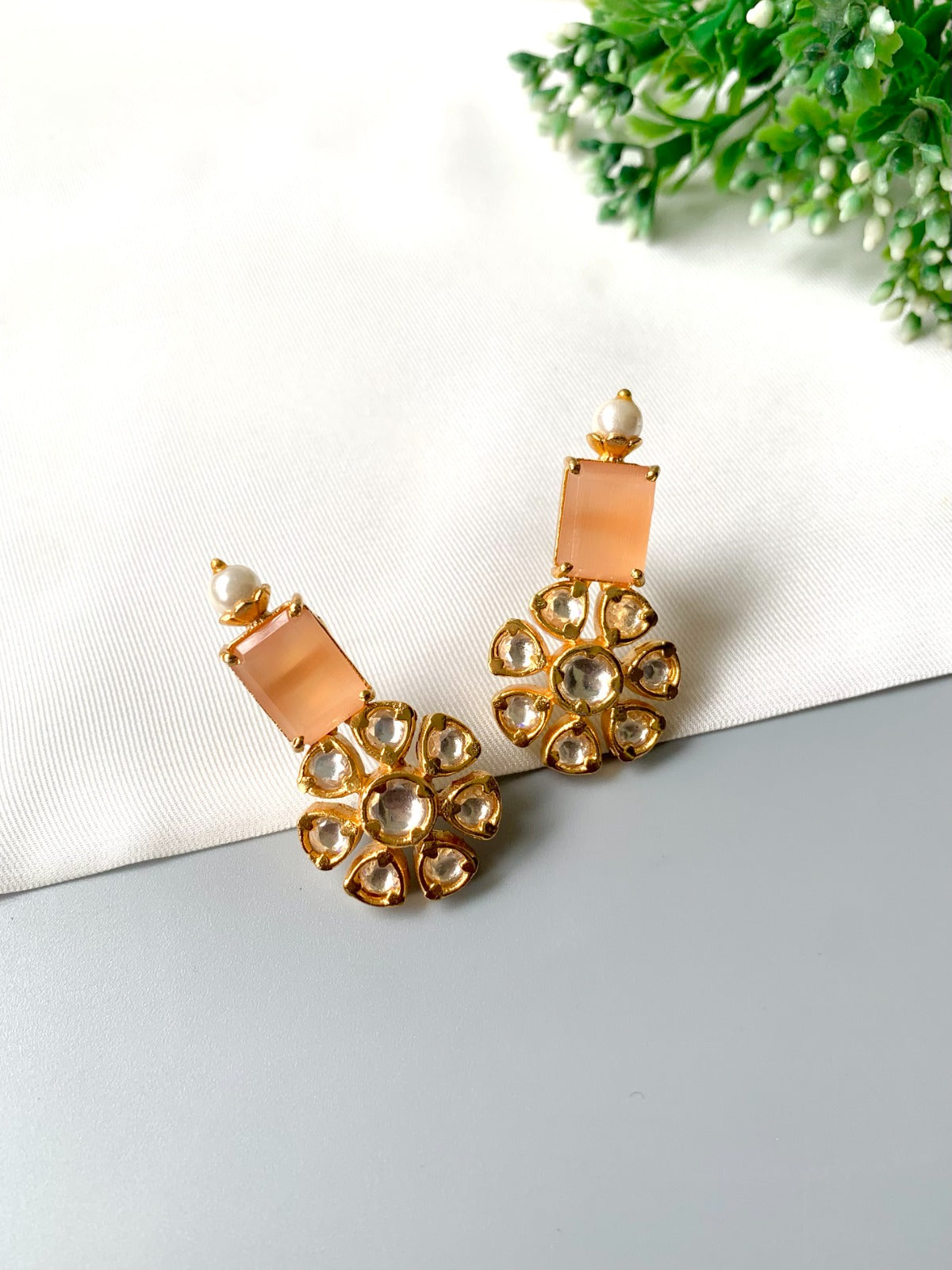 Pair of gold and peach-colored earrings on a white surface with greenery in the background