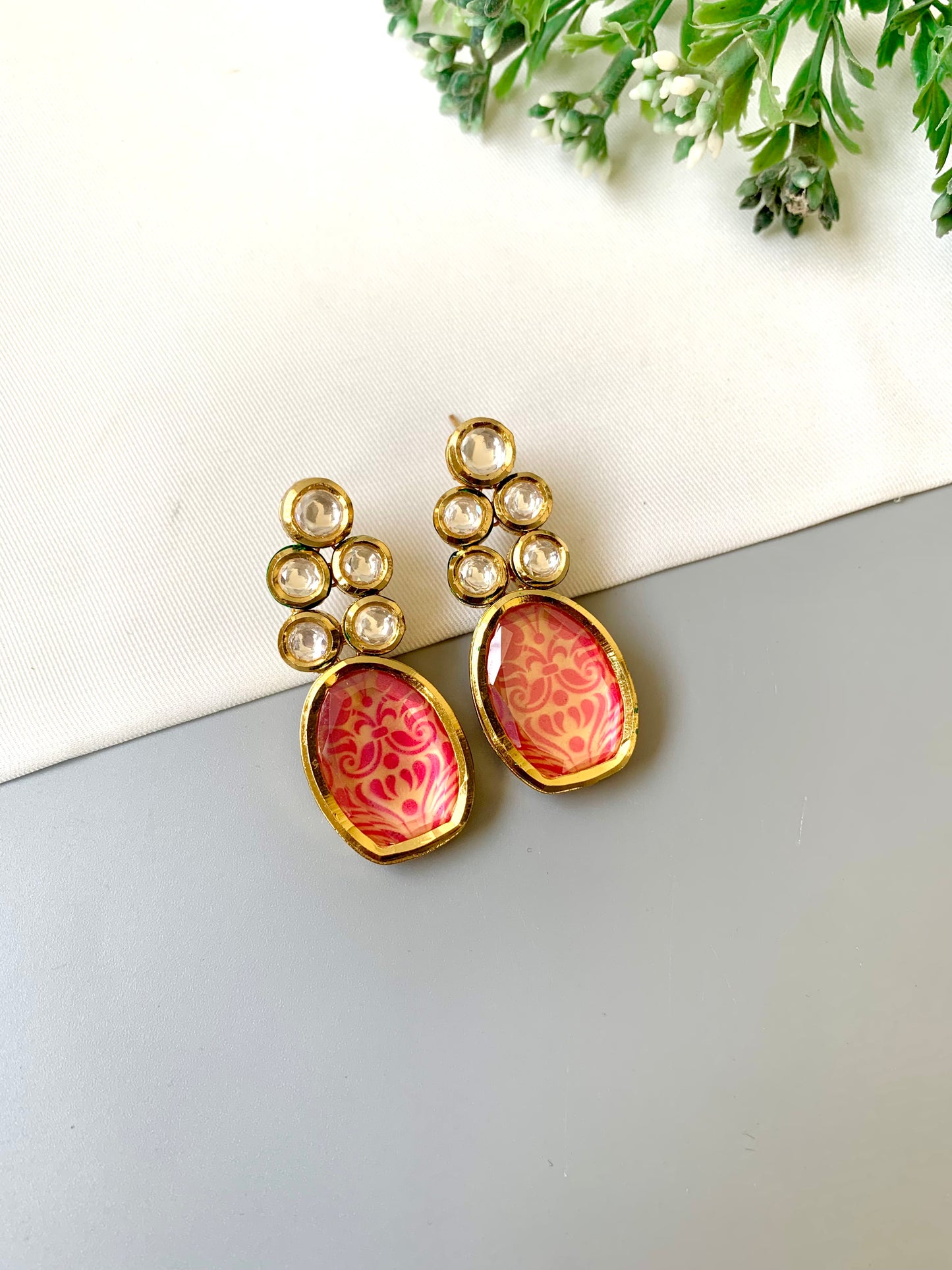 Gold earrings with pink floral designs on a light gray surface with greenery in the background