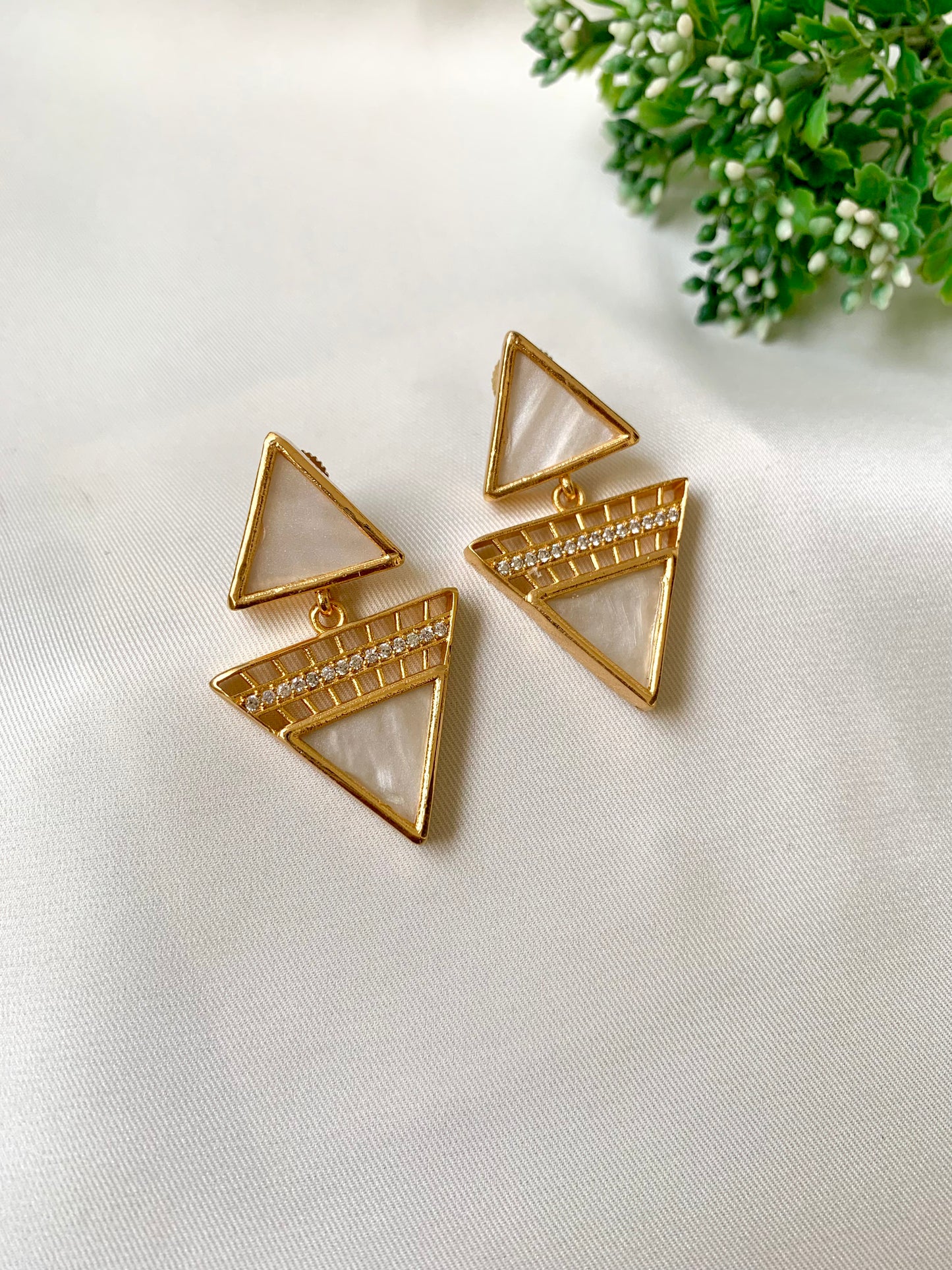 Gold triangle-shaped earrings on a white fabric background with greenery.
