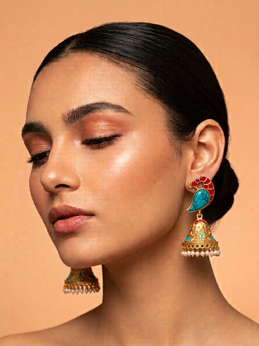 A close-up of a woman wearing ornate gold-toned jhumka earrings with turquoise blue, red, and green enamel details, set against a solid peach background.