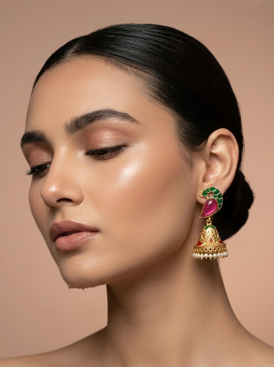 A close-up portrait of a woman wearing a traditional Indian jhumka earring. The earring features a gold-plated bell-shaped drop, a red stone top, and green stone accents, with small white stones along the bottom edge.