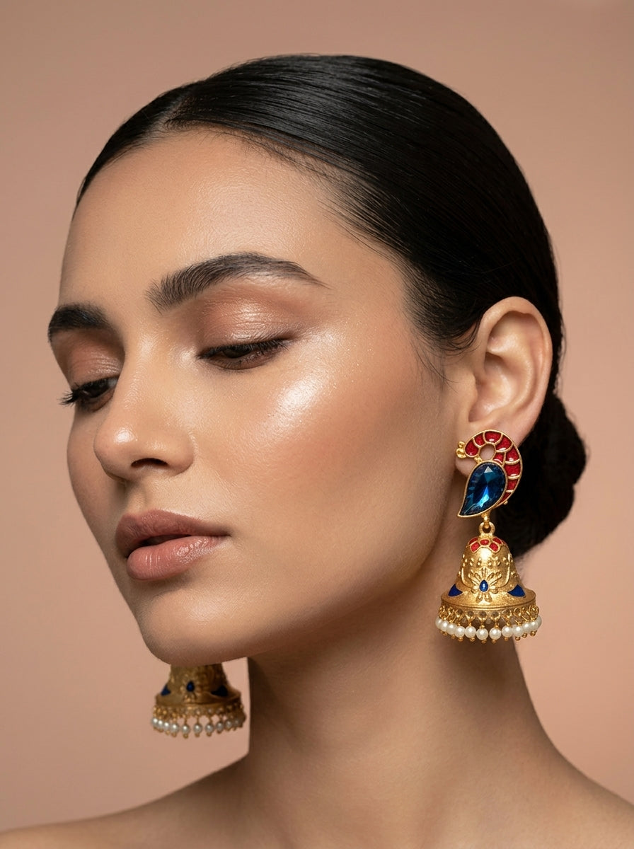 A close-up of a woman wearing ornate gold-toned jhumka earrings with red, blue, and white stone accents, against a soft beige background.