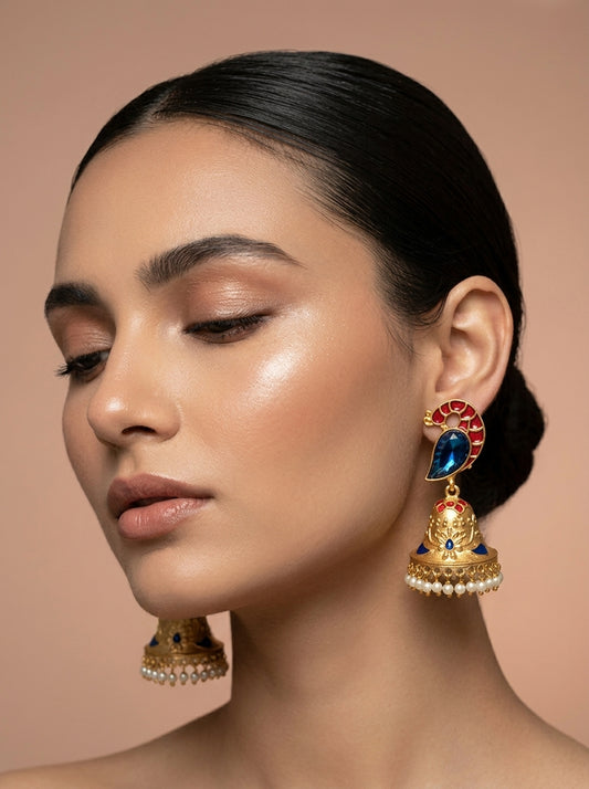 A close-up of a woman wearing ornate gold-toned jhumka earrings with red, blue, and white stone accents, against a soft beige background.