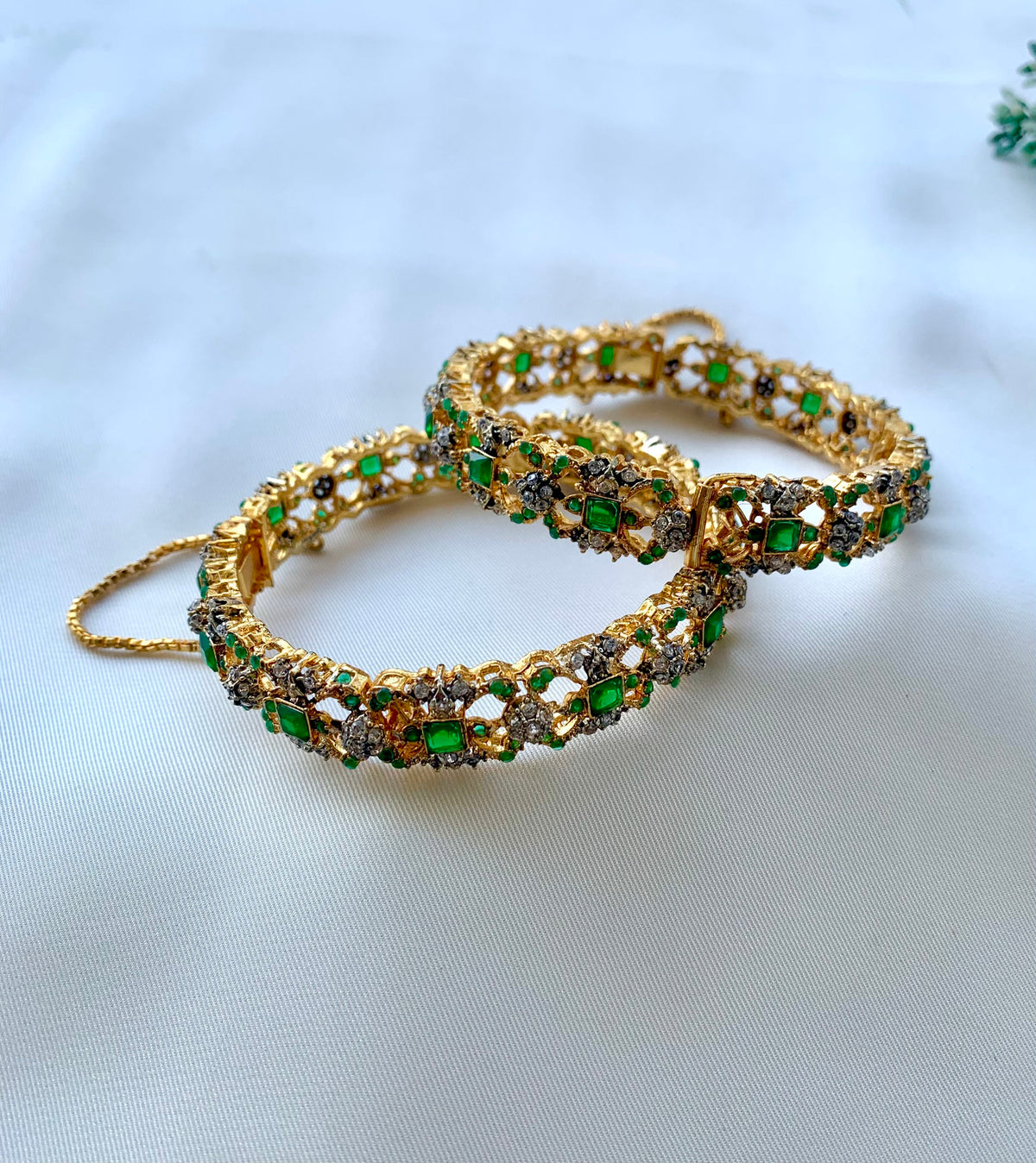 Two ornate gold-toned bangles decorated with green rectangular stones and small clear stones, arranged on a white fabric background.