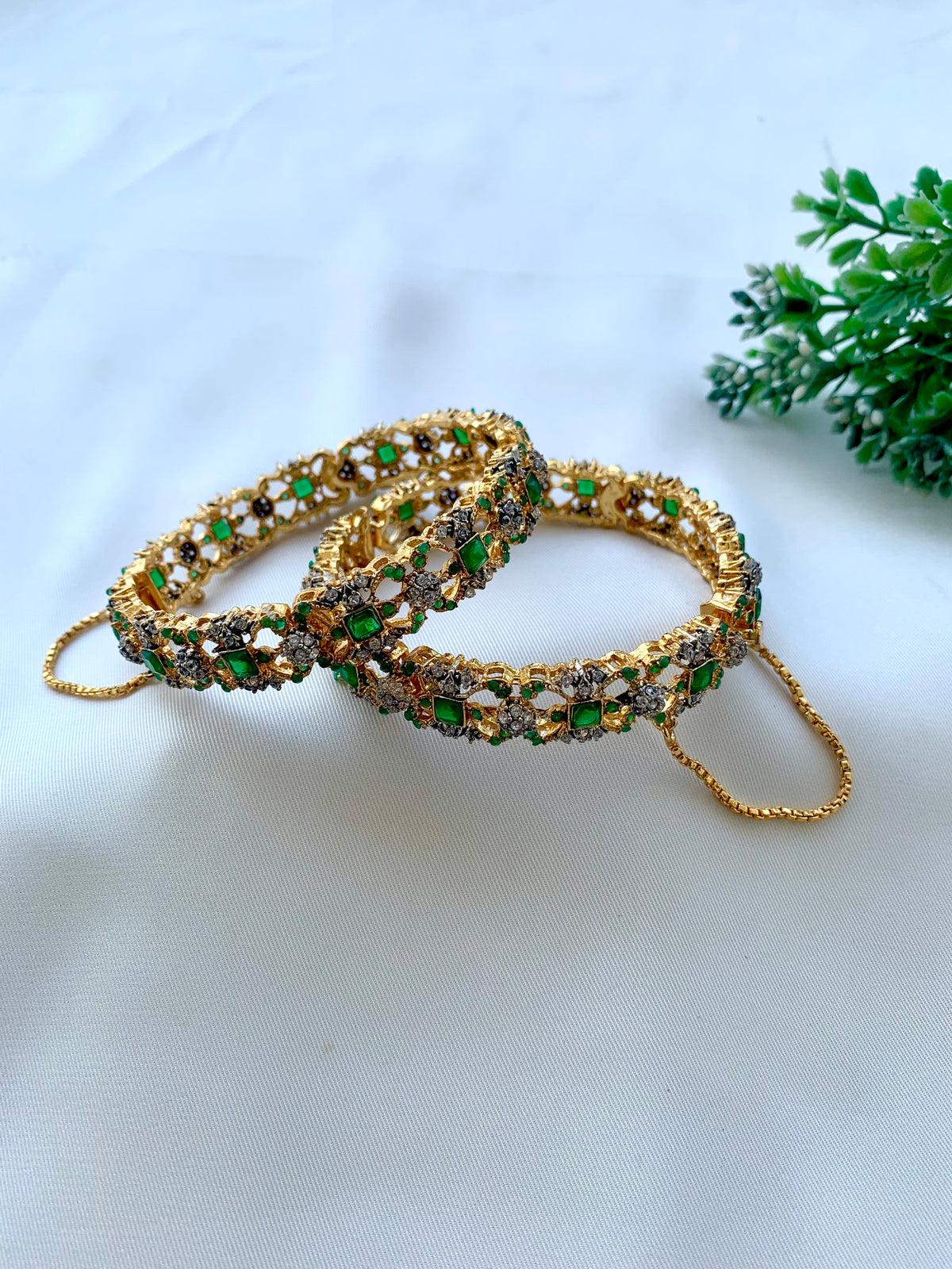 Two gold-plated bangles adorned with green and brown stones, displayed on a white fabric background with a small green plant in the corner.