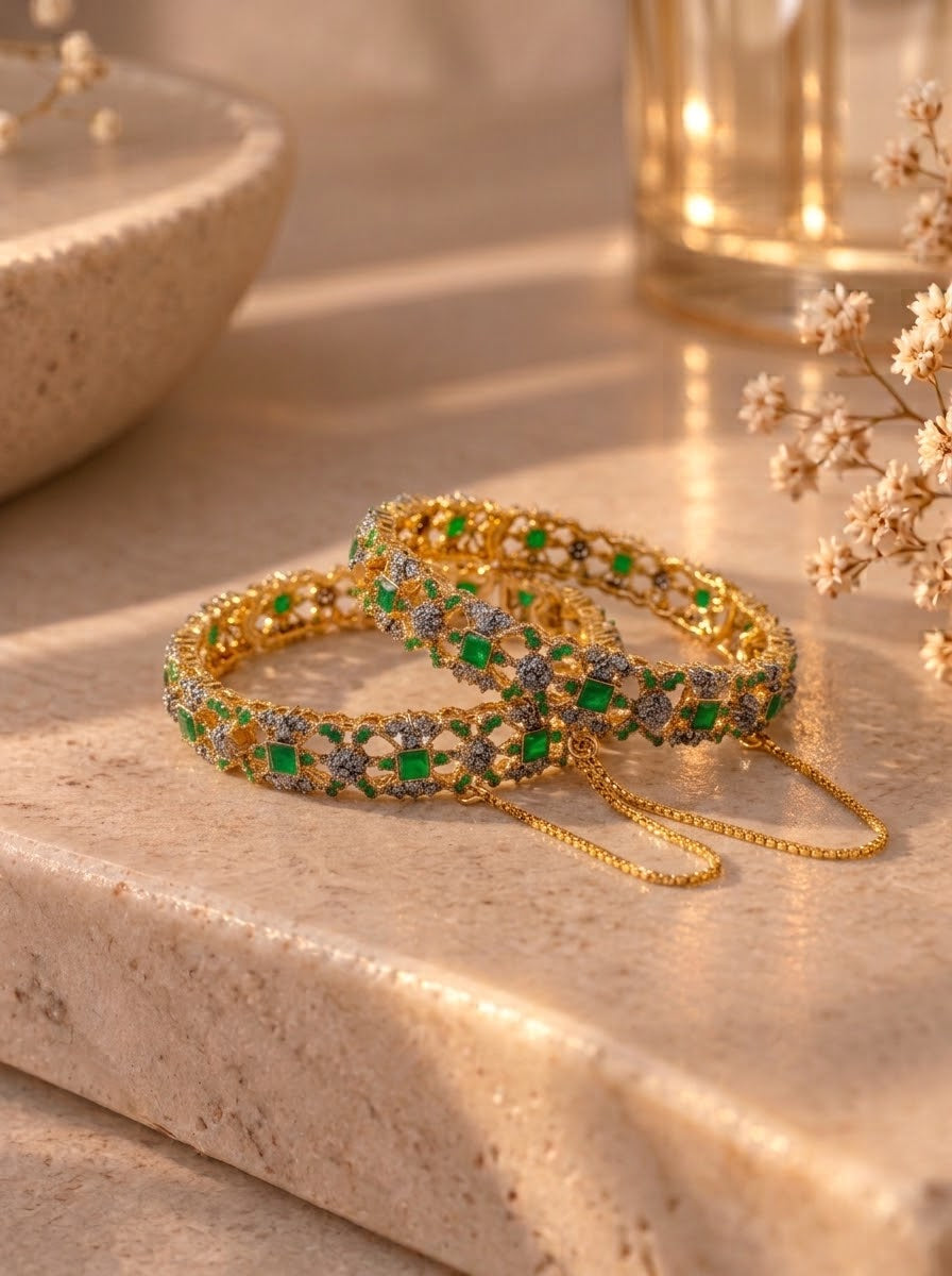 Three gold-plated bangles adorned with green and silver stones, arranged on a beige stone surface with soft warm lighting and dried flowers in the background.
