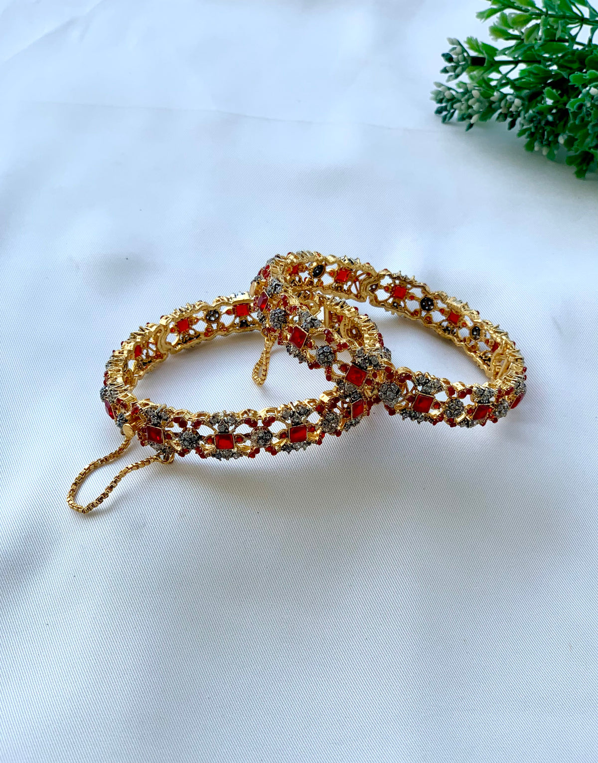 Two ornate gold-toned bangles decorated with red stones and small clear stones, arranged on a white fabric surface with a small green plant in the corner.