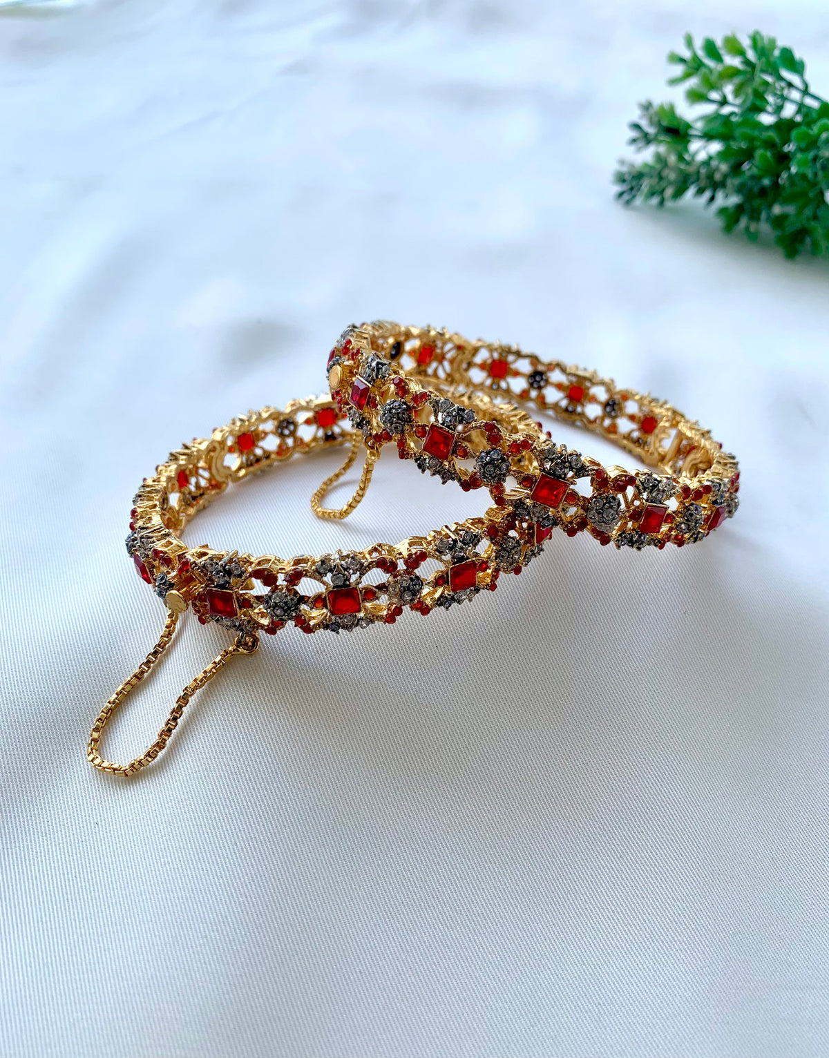 A pair of ornate gold-toned bangles decorated with red and silver stones, displayed on a white fabric background with a small green plant in the corner.