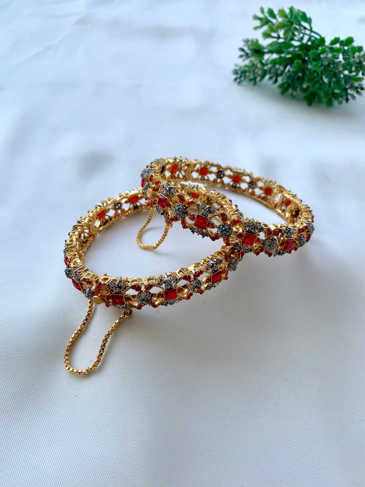Two ornate gold-toned bangles decorated with red and silver stones, displayed on a white fabric background with a small green plant in the corner.