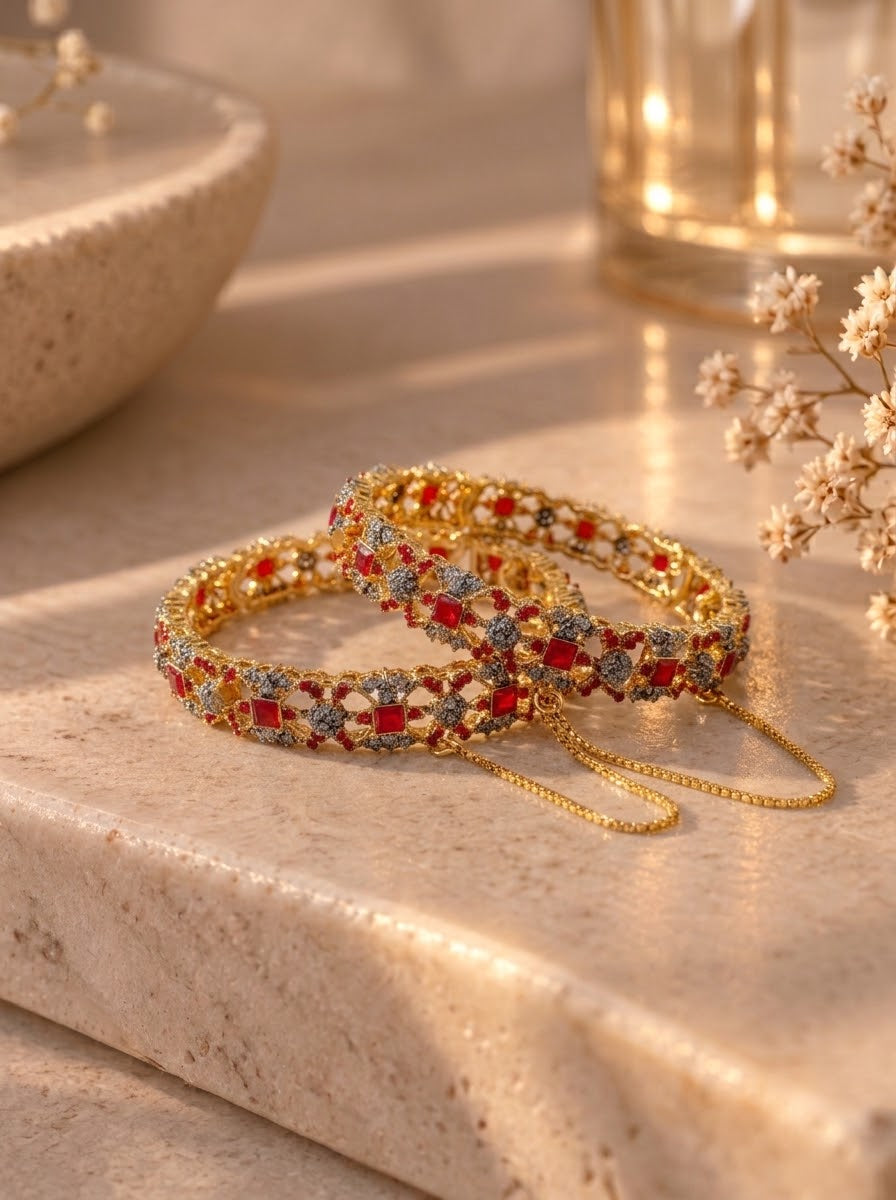 Two gold-plated bangles adorned with red and blue stones, resting on a beige stone surface with warm, soft-focus background elements.