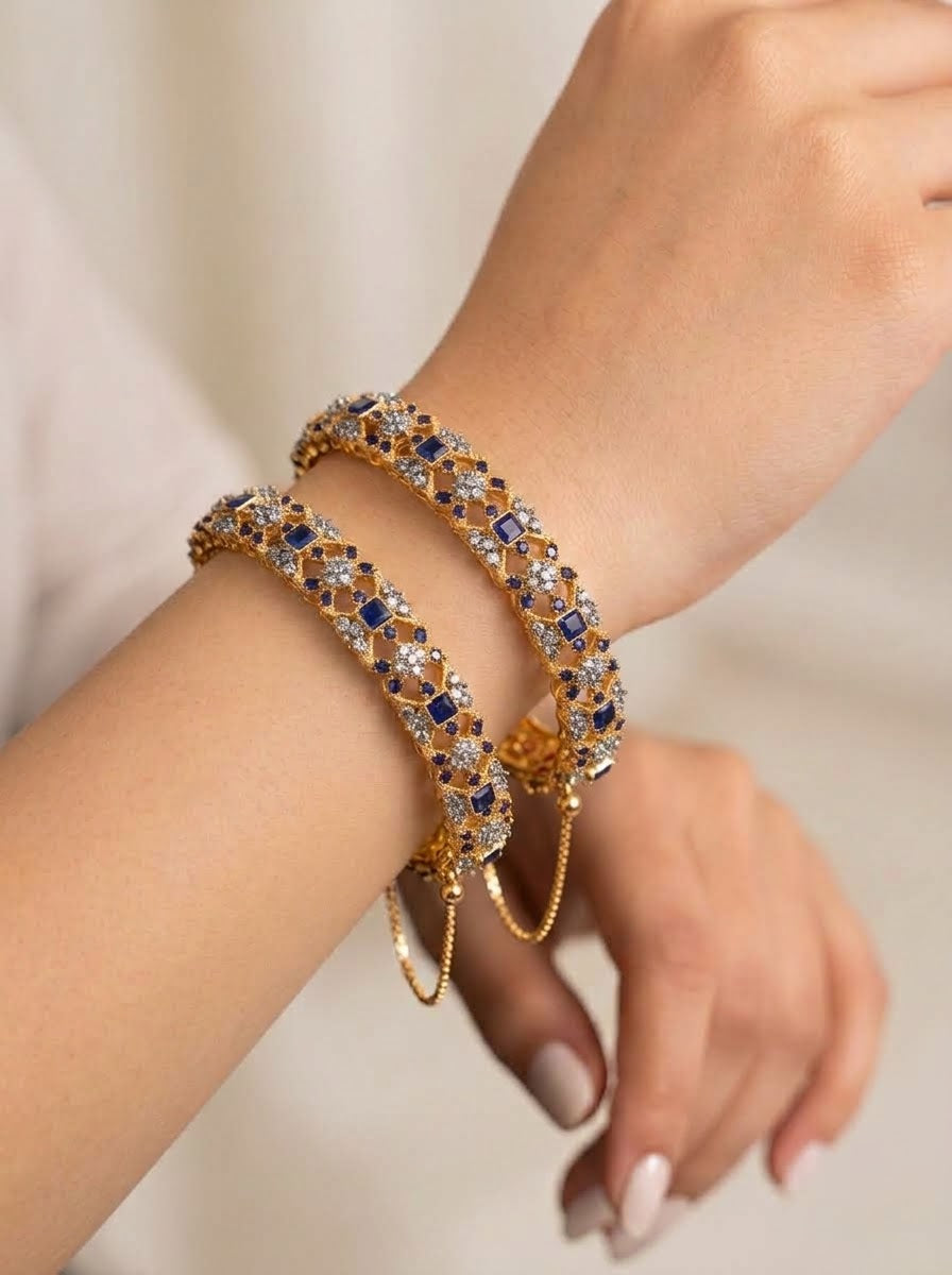 A close-up of a person's wrist wearing two gold-toned bangle bracelets adorned with blue, white, and gold crystals.