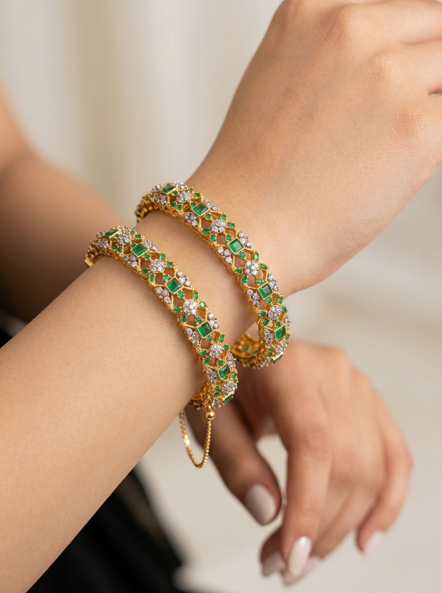 A close-up of a person's wrist wearing two stacked gold bangle bracelets decorated with green and white stones.