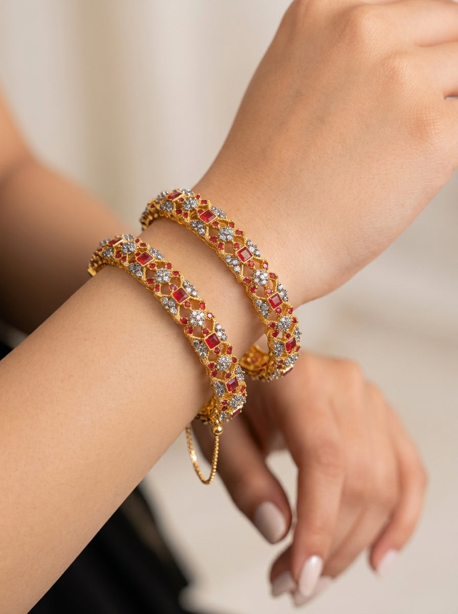 A close-up of a person's wrist wearing two interlocking gold-toned bangle bracelets adorned with red and clear rhinestones, against a soft beige background.
