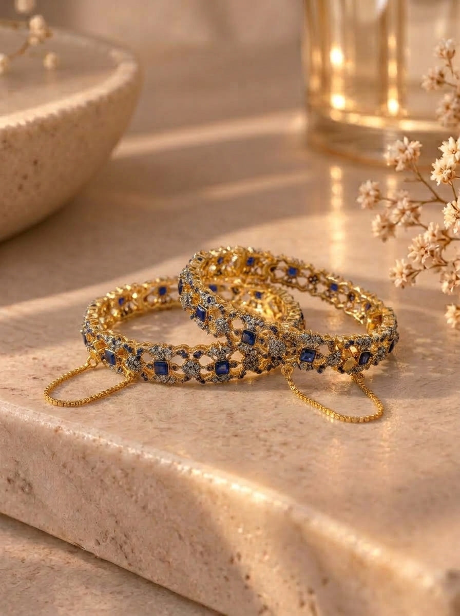 Two gold-toned bangles with blue and clear stones, displayed on a beige stone surface with decorative dried flowers and a softly lit background.