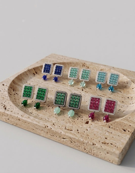 Six pairs of stud earrings arranged on a beige travertine tray against a light gray background. The earrings feature square grid patterns in blue, green, aqua, and magenta, with small round beads in matching colors below each pair.