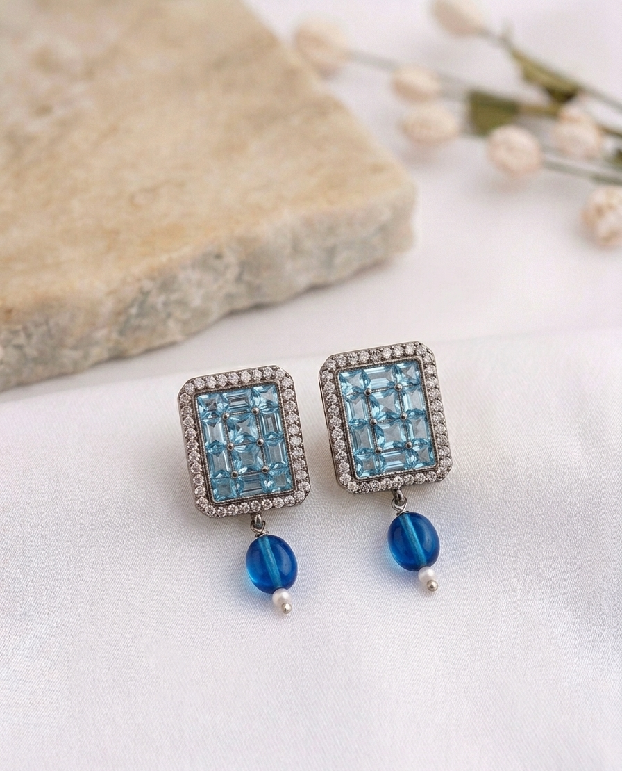 A pair of rectangular drop earrings featuring light blue emerald-cut stones surrounded by small clear stones, with blue bead drops and silver-toned settings, displayed on a white fabric surface with a stone and dried flowers in the background.