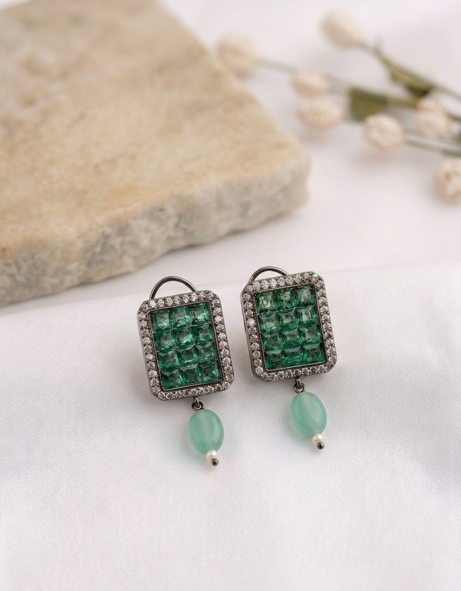 A pair of rectangular drop earrings with green faceted gemstone centers, silver-toned crystal-encrusted borders, and dangling mint-green beads, displayed on a white fabric surface with a stone prop.