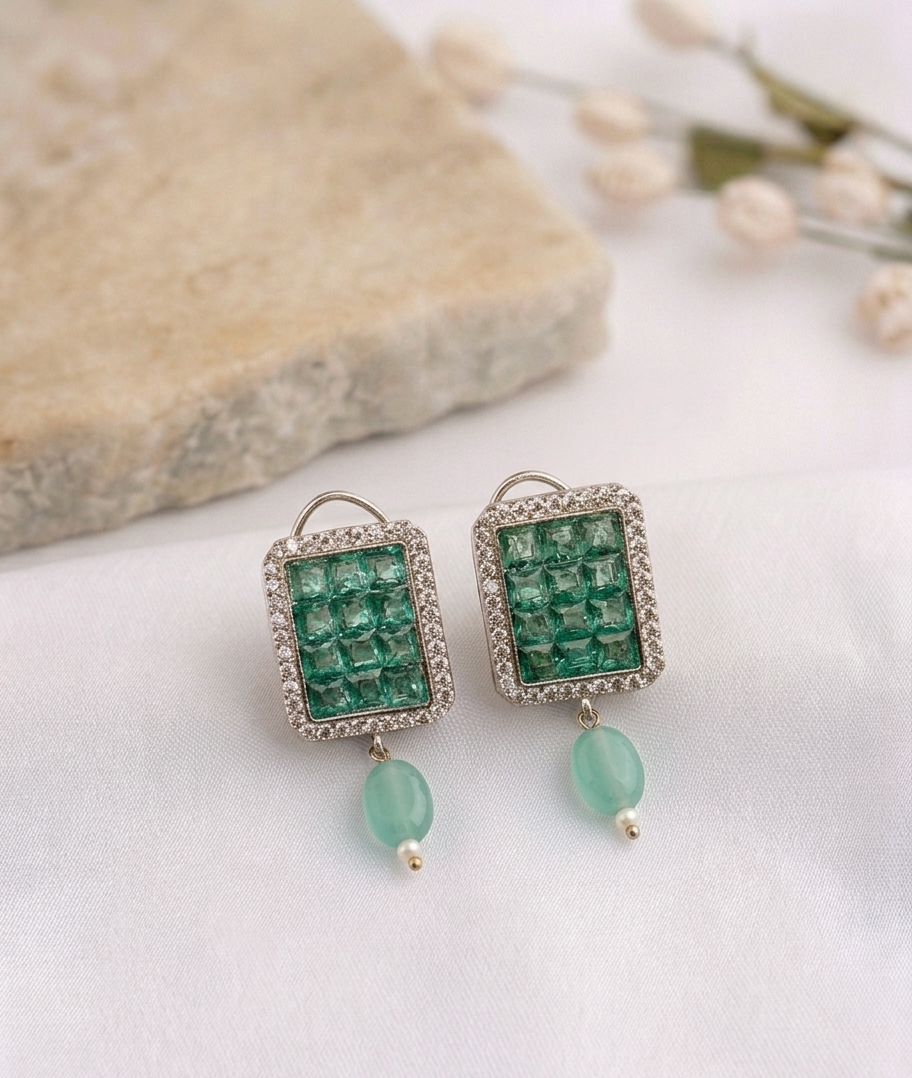 A pair of rectangular drop earrings with emerald-cut green stones in a silver-toned setting, accented with small clear stones and a dangling light green bead, displayed on a white fabric surface with a stone and dried flowers in the background.