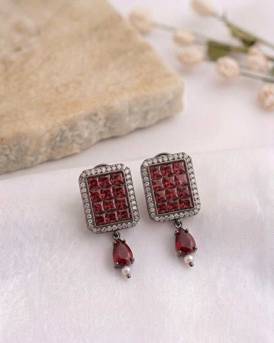 A pair of rectangular drop earrings with deep red faceted gemstones in a silver-toned setting, decorated with small clear stones and hanging teardrop red gemstones with tiny pearl-like accents, displayed on a white textured surface with a stone and dried flowers in the background.