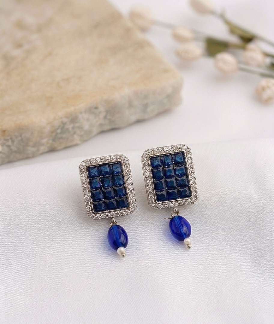 A pair of rectangular drop earrings with deep blue faceted stones in a textured grid, surrounded by a silver-tone crystal-encrusted border, with a dangling blue bead and small pearl accent below, displayed on a white fabric surface with a stone and dried flowers in the background.