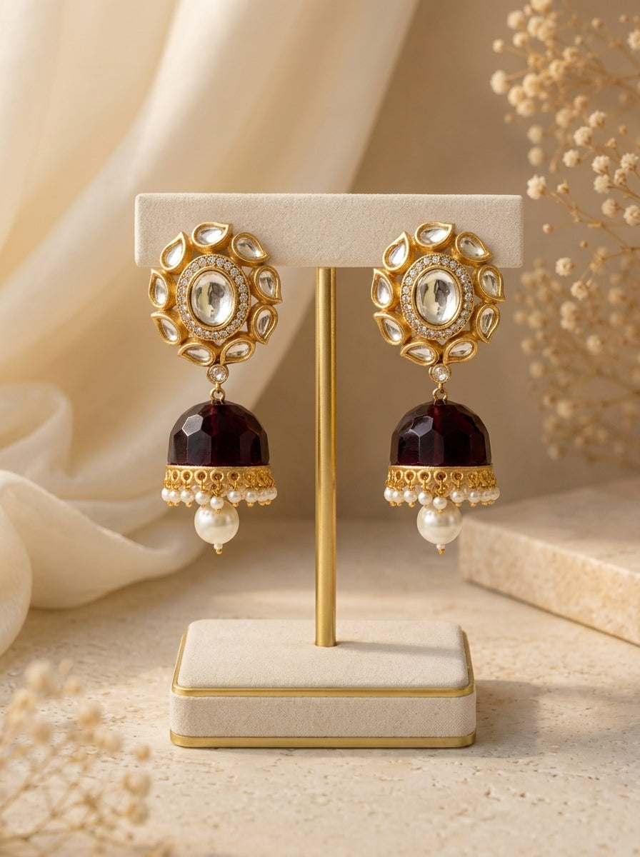 A pair of ornate gold-toned jhumka earrings displayed on a cream jewelry stand. Each earring features a circular gold frame with clear stones, a dark red dangling bell, and a small white pearl at the bottom, set against a soft beige background with dried floral decor.