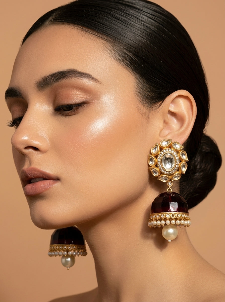 A close-up of a woman wearing a pair of ornate gold-plated jhumka earrings with dark red bell-shaped stones, white faux pearls, and clear rhinestones, set against a solid warm beige background.