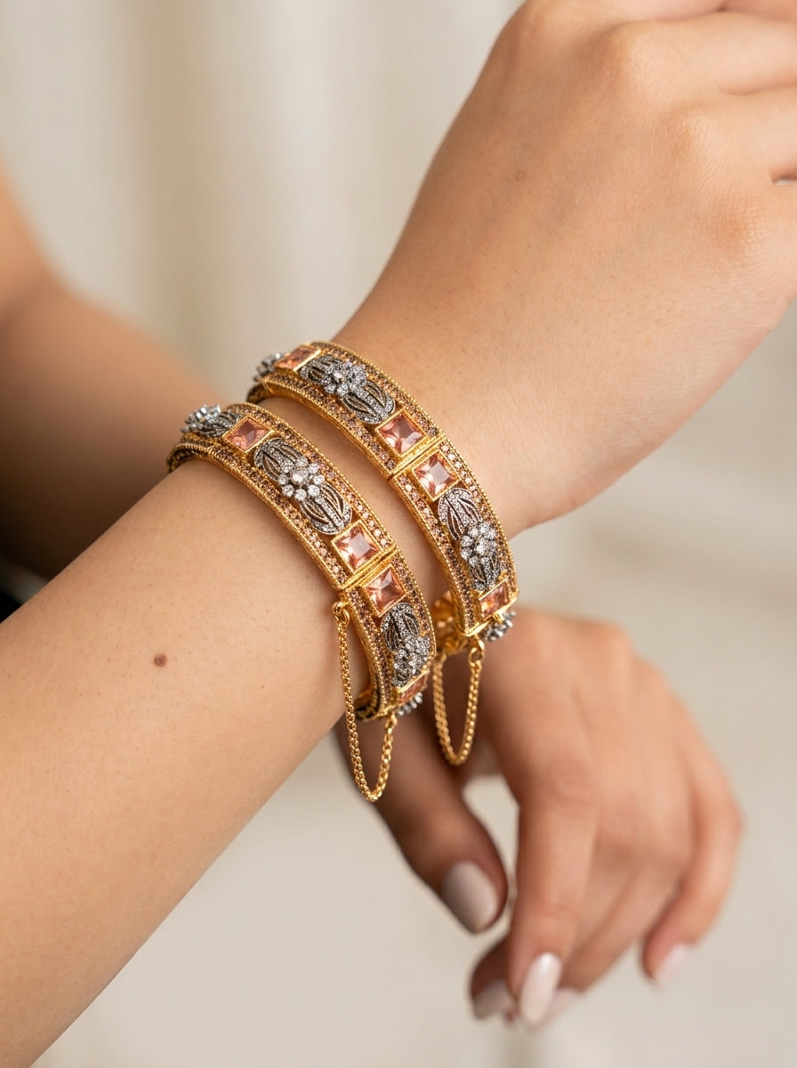 A close-up of a person's wrist wearing two ornate gold bangles decorated with silver-toned floral motifs and pink stones, against a soft neutral background.