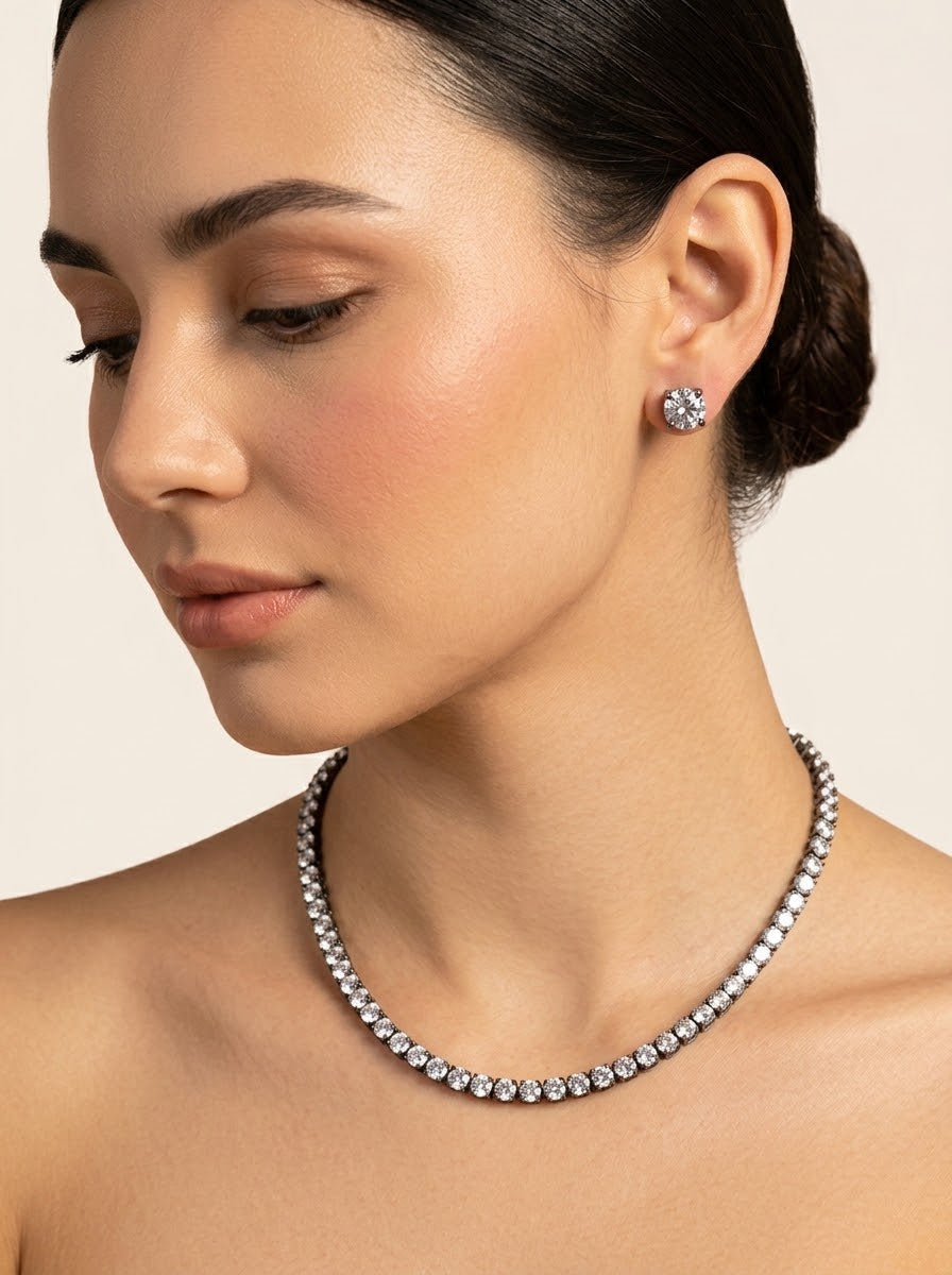 A close-up of a woman wearing a delicate silver tennis necklace and a matching stud earring. The jewelry features clear gemstones set in silver-toned metal, shown against a soft beige background.