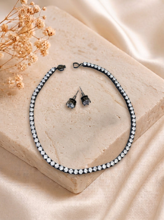 A black chain necklace and matching stud earrings set with clear rhinestones, arranged on a beige stone block with dried flowers and cream satin fabric in the background.