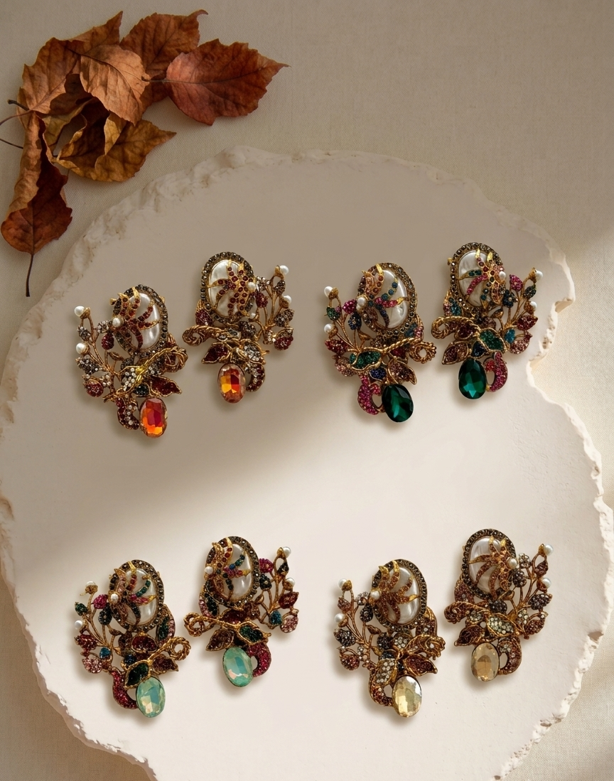 Six ornate vintage-style brooches with carved cameo motifs and colorful rhinestone accents, arranged on a white ceramic plate with dried autumn leaves in the corner.