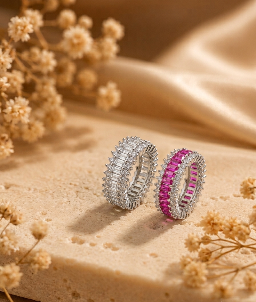 Two silver rings with rectangular gemstones, one silver and one pink, displayed on a beige stone surface with dried flowers and draped satin fabric.